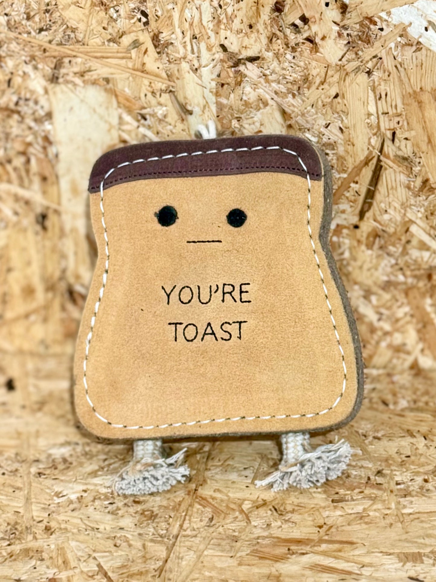 A brown jute toy in the shape of a stylized bread loaf with a smiling face and the text 'YOU'RE TOAST' printed on it, featuring suede accents and a rope handle.