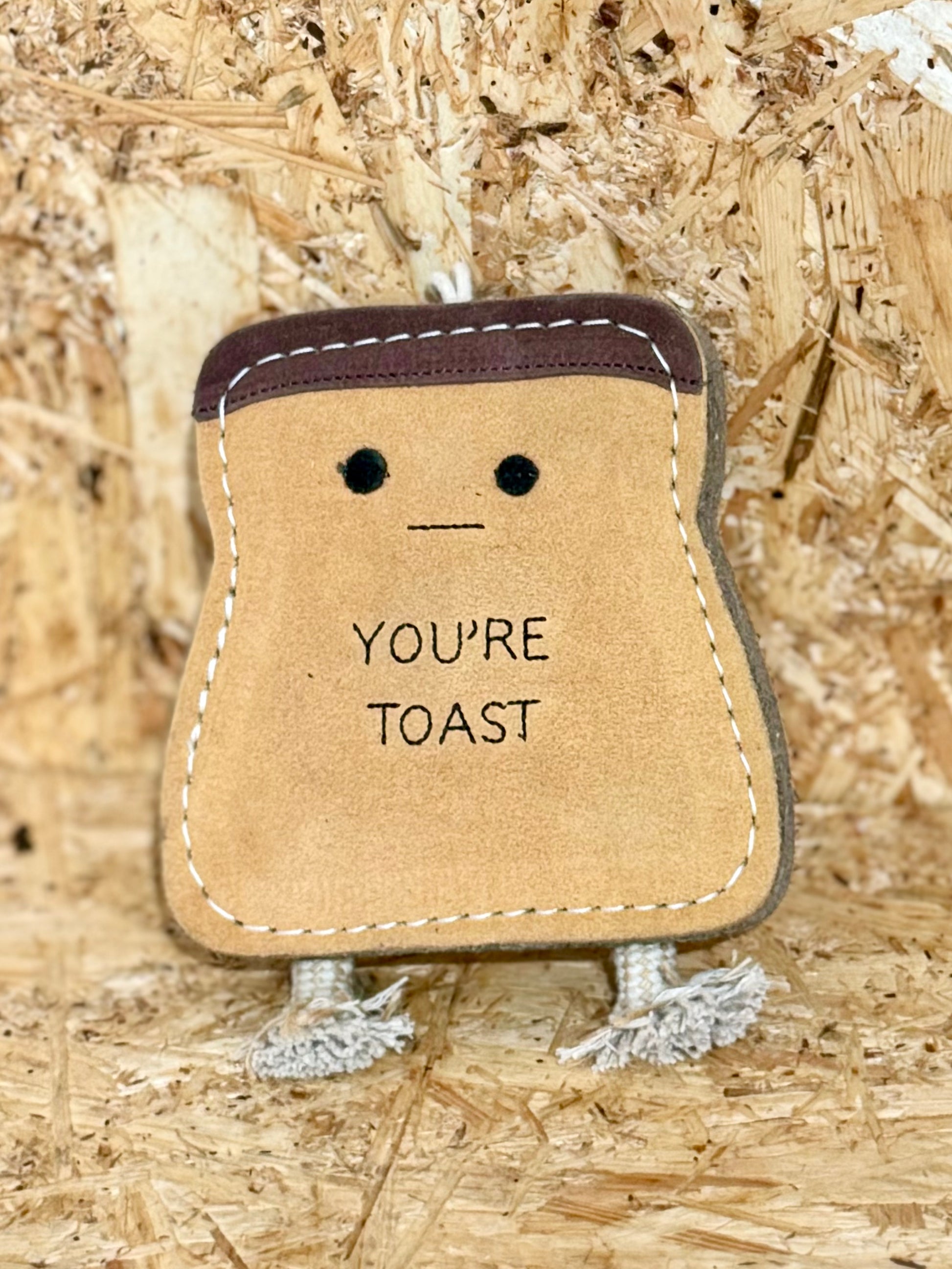 A brown jute toy in the shape of a stylized bread loaf with a smiling face and the text 'YOU'RE TOAST' printed on it, featuring suede accents and a rope handle.