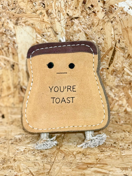 A brown jute toy in the shape of a stylized bread loaf with a smiling face and the text 'YOU'RE TOAST' printed on it, featuring suede accents and a rope handle.
